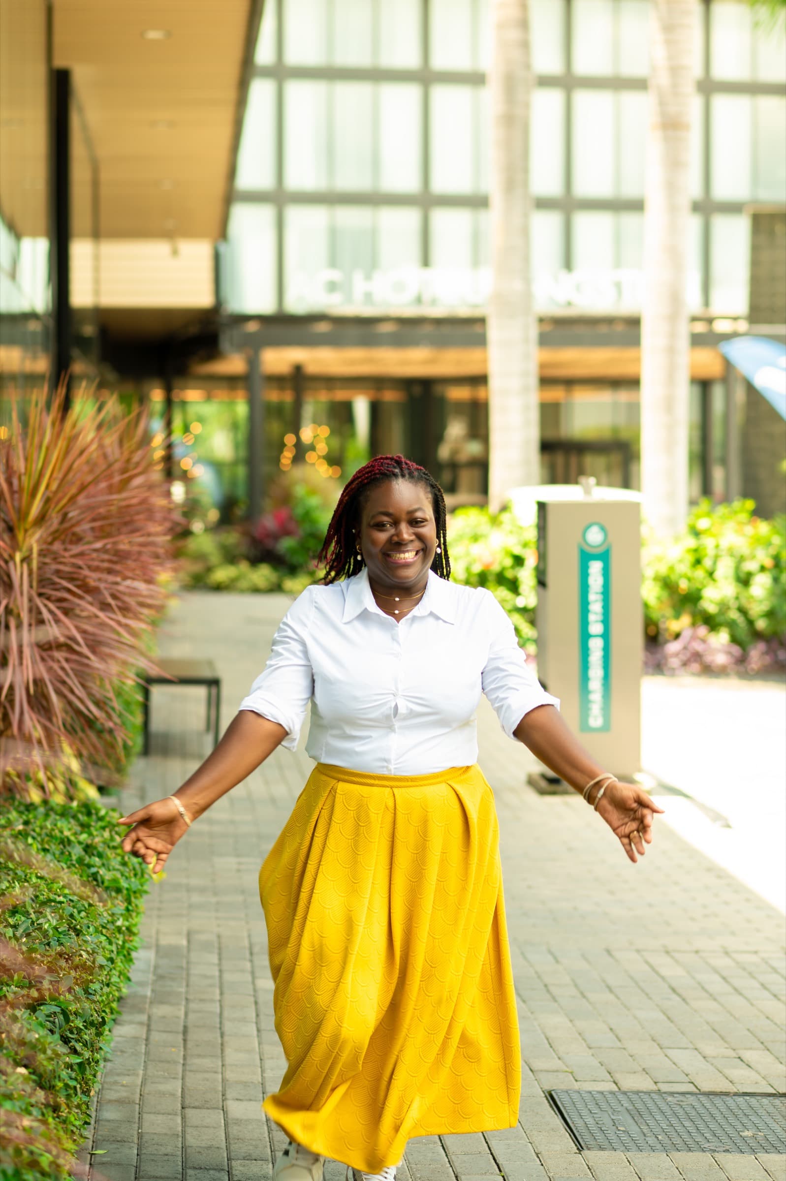 Anwin Mbah walking outdoors in a yellow skirt, smiling, mid-stride against a sunlit modern building